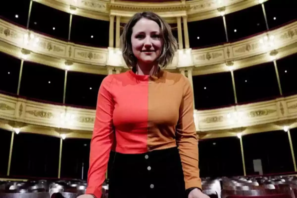 Short-haired, wavy-haired woman wearing a two-toned blouse, red and orange, standing inside an elegant theater with illuminated balconies. Short-haired, wavy-haired woman wearing a two-toned blouse, red and orange, standing inside an elegant theater with illuminated balconies.