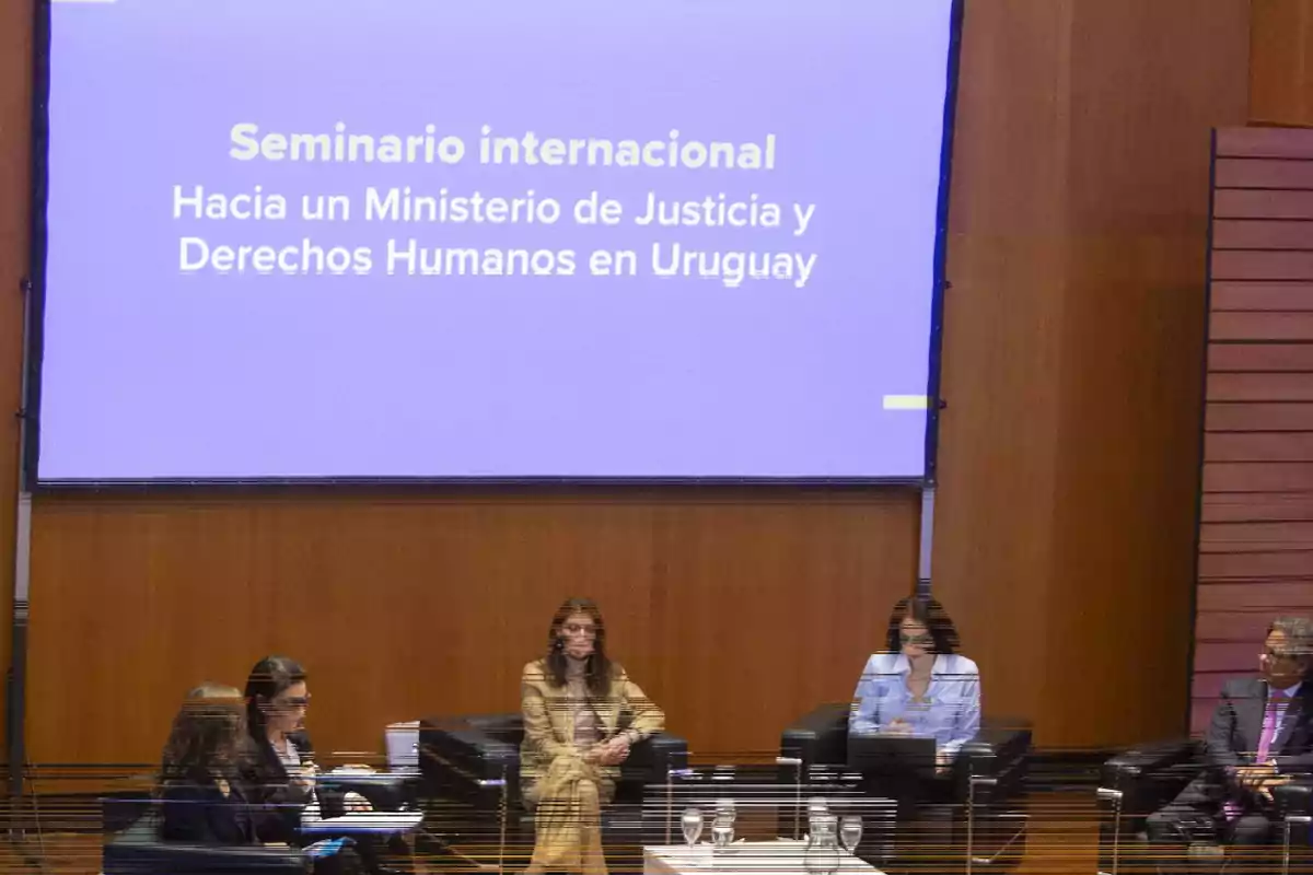 Five people seated in black armchairs participate in a panel in front of a screen displaying the title International Seminar: Towards a Ministry of Justice and Human Rights in Uruguay. Five people seated in black armchairs participate in a panel in front of a screen displaying the title International Seminar: Towards a Ministry of Justice and Human Rights in Uruguay.