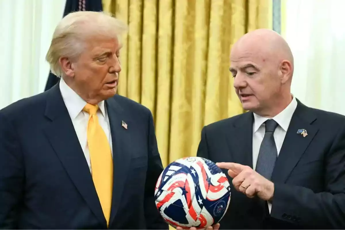 Dos hombres de traje conversan mientras uno de ellos sostiene un balón de fútbol con diseño de la bandera de Estados Unidos frente a una cortina dorada Dos hombres de traje conversan mientras uno de ellos sostiene un balón de fútbol con diseño de la bandera de Estados Unidos frente a una cortina dorada