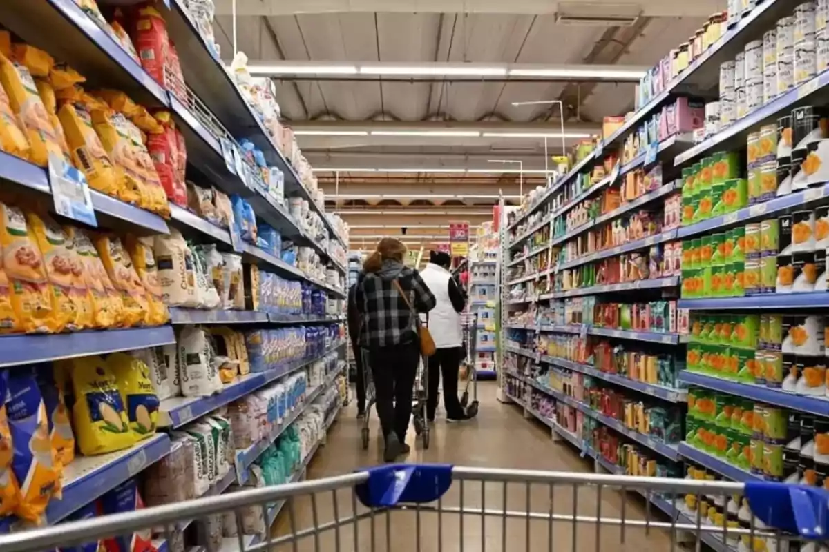 Pasillo de supermercado con estantes llenos de productos y personas empujando carritos de compras