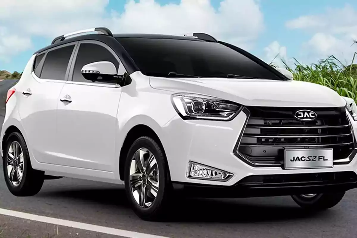 Auto blanco JAC S2 FL estacionado en una carretera con vegetación al fondo y cielo parcialmente nublado Auto blanco JAC S2 FL estacionado en una carretera con vegetación al fondo y cielo parcialmente nublado