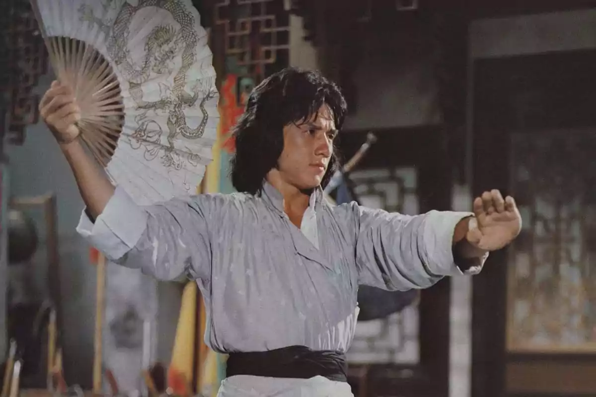 Young man with long hair dressed in traditional Chinese clothing holding a fan decorated with a dragon while striking a martial arts pose in an ancient interior Young man with long hair dressed in traditional Chinese clothing holding a fan decorated with a dragon while striking a martial arts pose in an ancient interior