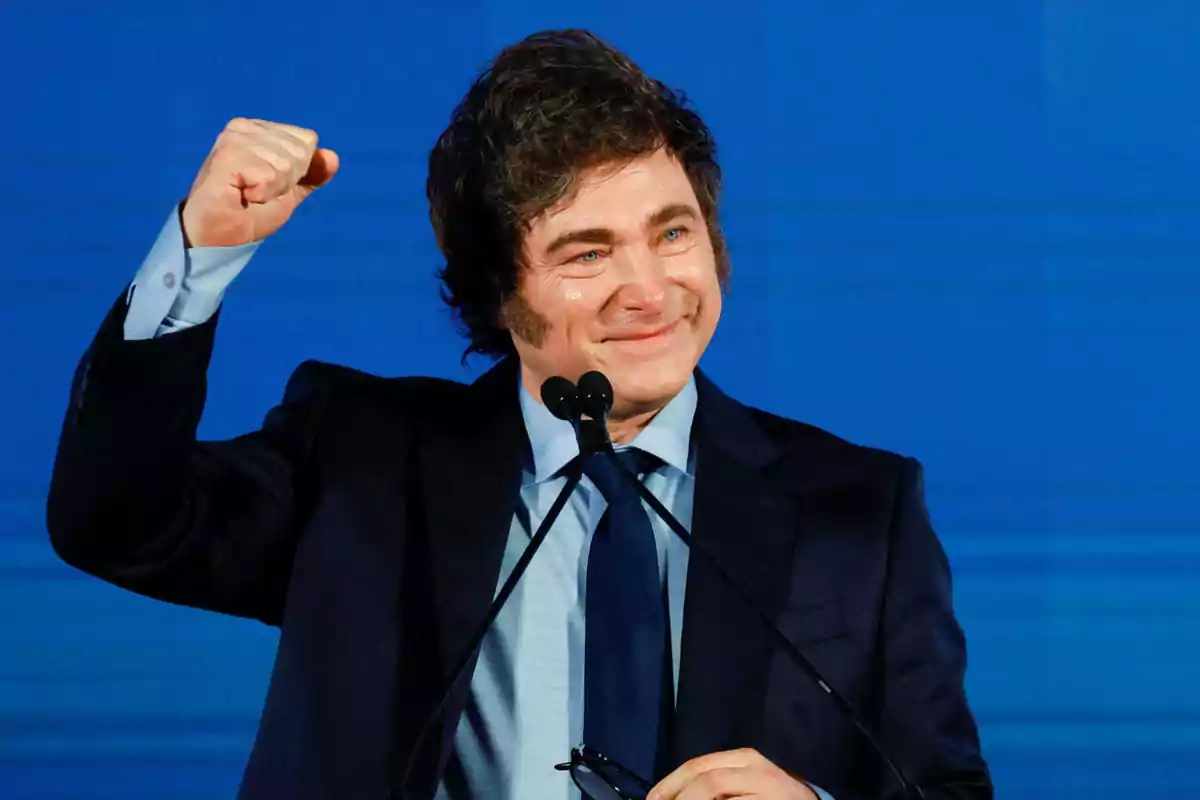 Dark-haired man in a blue suit smiling and raising his fist in front of a microphone and a blue background Dark-haired man in a blue suit smiling and raising his fist in front of a microphone and a blue background