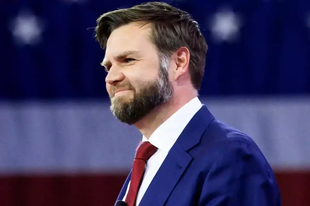 Man with a beard and short hair wearing a blue suit and red tie in front of a blurred United States flag Man with a beard and short hair wearing a blue suit and red tie in front of a blurred United States flag