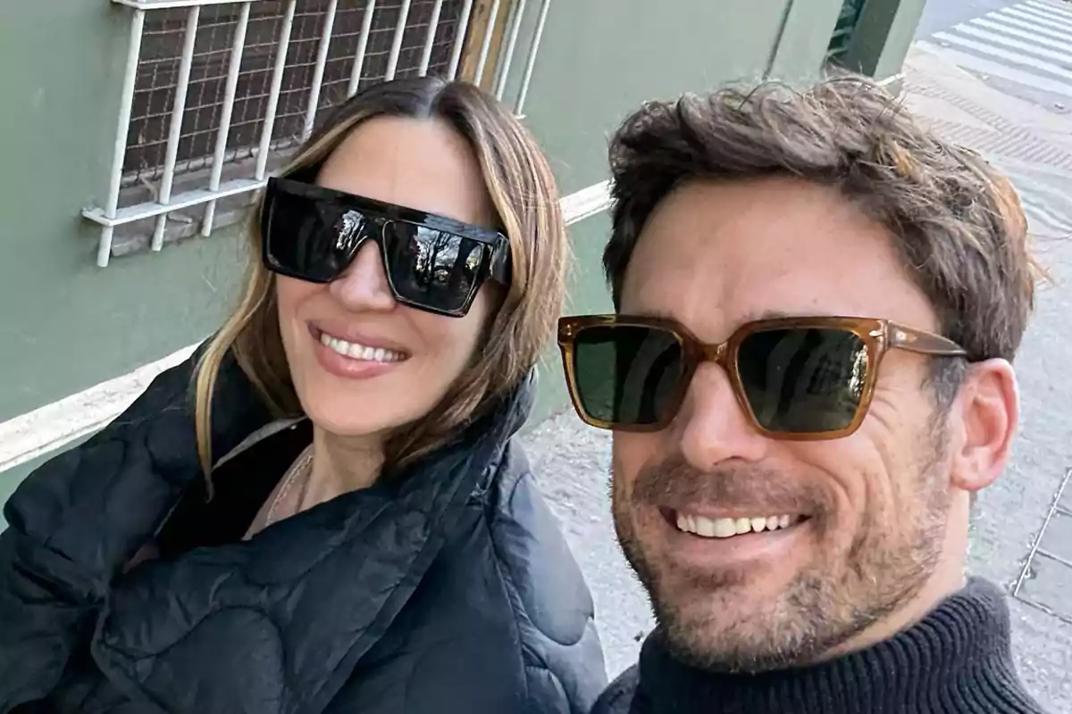 Dos personas sonríen usando gafas de sol mientras se toman una selfie en la calle.