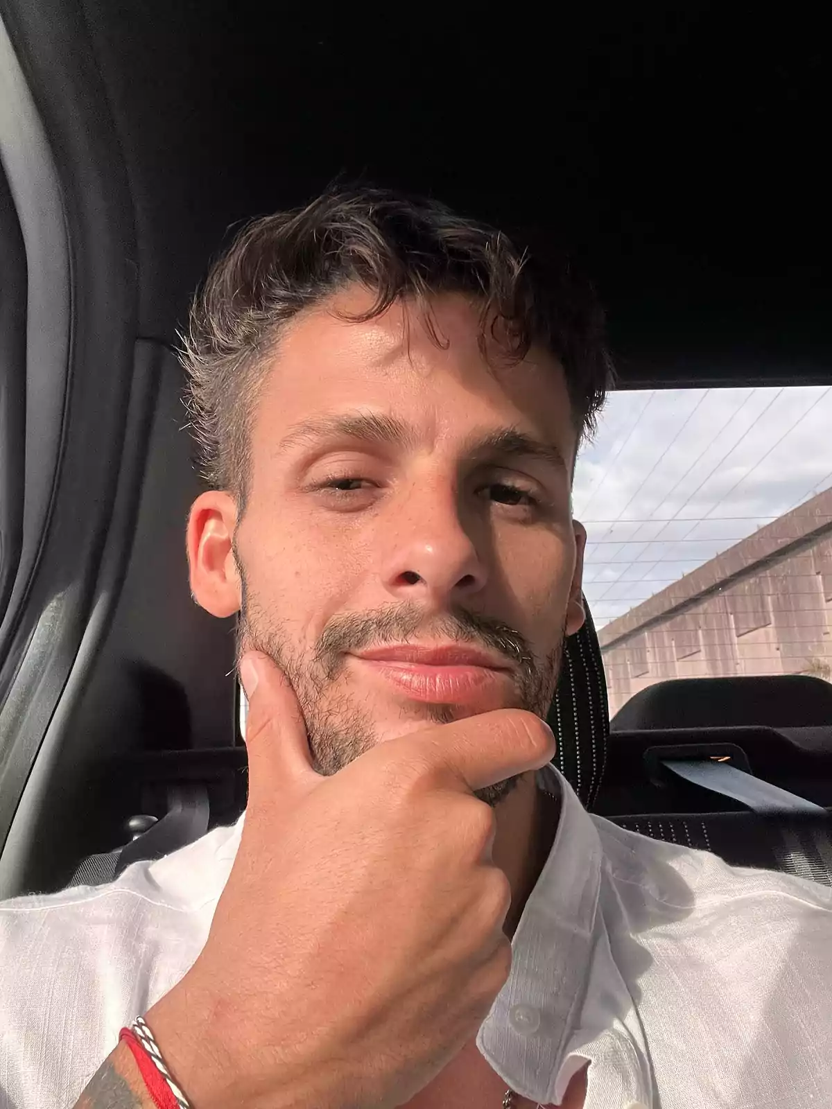 Hombre joven con barba corta y cabello ondulado sentado en el asiento trasero de un auto, usando una camisa blanca y posando con la mano en el mentón mientras mira a la cámara Hombre joven con barba corta y cabello ondulado sentado en el asiento trasero de un auto, usando una camisa blanca y posando con la mano en el mentón mientras mira a la cámara