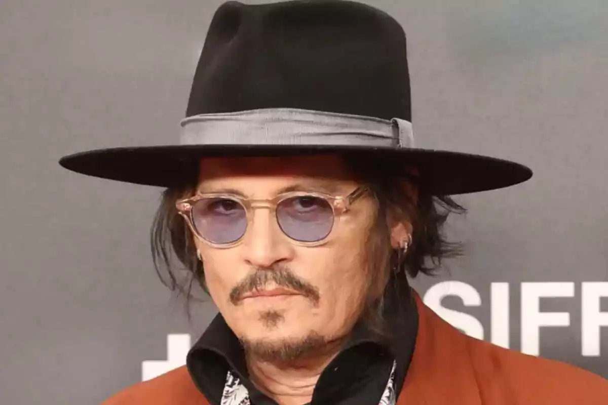 Hombre con sombrero negro, gafas de sol redondas y chaqueta marrón en un evento Hombre con sombrero negro, gafas de sol redondas y chaqueta marrón en un evento