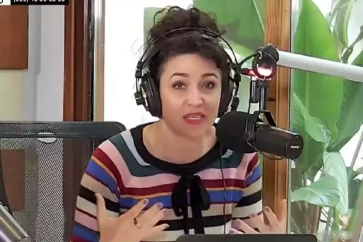 Mujer con audífonos y micrófono hablando en una cabina de radio, usando un suéter de rayas de colores y gesticulando con las manos.