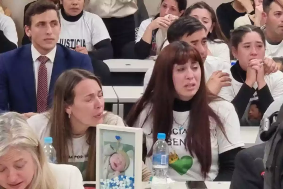 Varias personas lloran y muestran expresiones de dolor en una sala, algunas llevan camisetas blancas con mensajes de justicia y en la mesa hay una foto de un bebé enmarcada junto a una botella de agua Varias personas lloran y muestran expresiones de dolor en una sala, algunas llevan camisetas blancas con mensajes de justicia y en la mesa hay una foto de un bebé enmarcada junto a una botella de agua