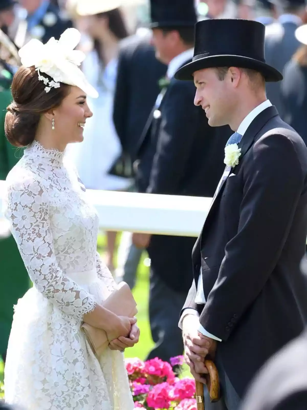 Una pareja elegantemente vestida sonríe y se mira de frente en un evento al aire libre rodeados de flores y otras personas en el fondo Una pareja elegantemente vestida sonríe y se mira de frente en un evento al aire libre rodeados de flores y otras personas en el fondo