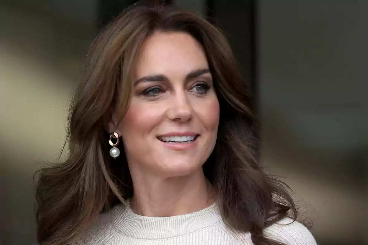 Mujer de cabello castaño largo y suelto con suéter claro y aretes de perla sonríe mirando hacia un lado