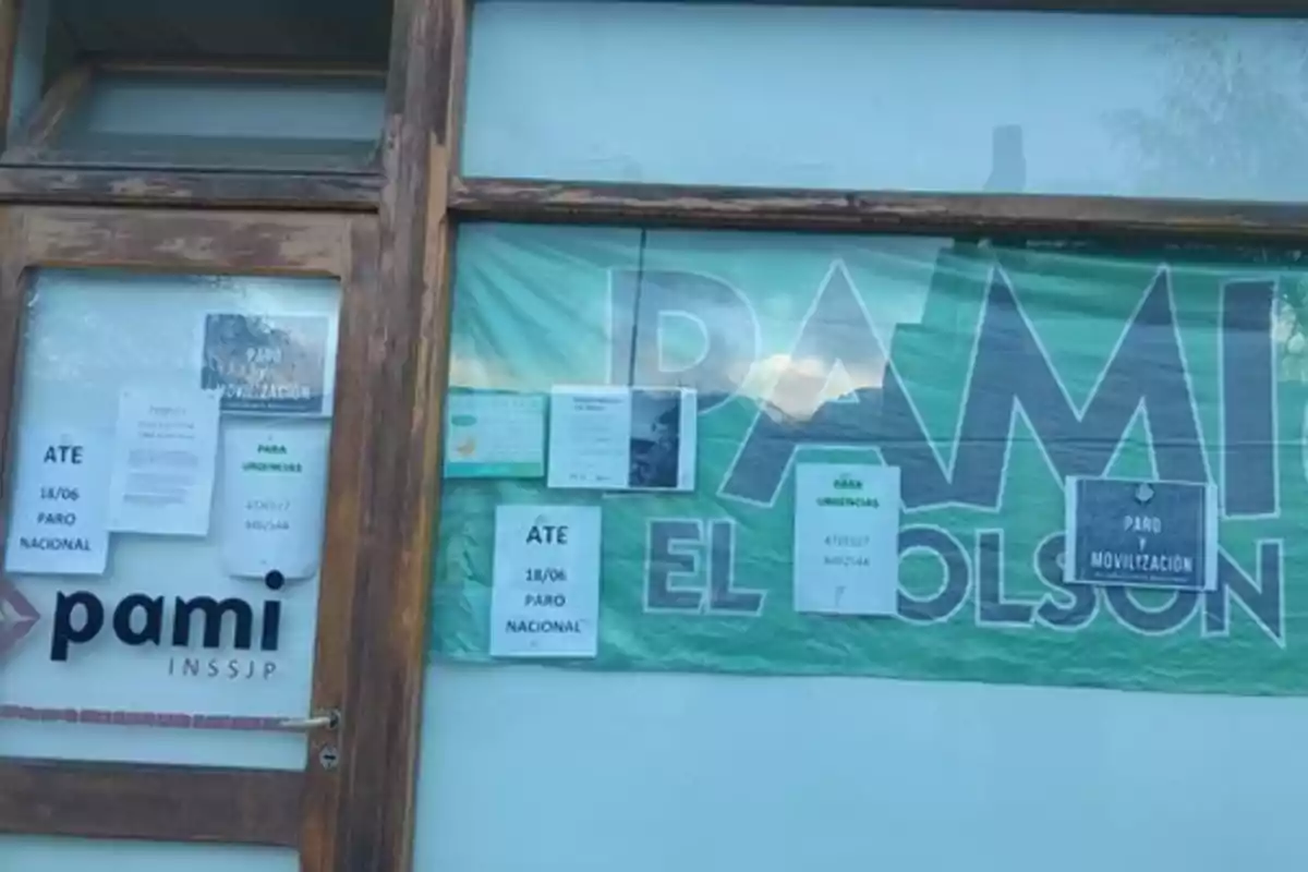 Fachada de una oficina de PAMI con varios carteles pegados en la puerta y ventana anunciando paro nacional y números para urgencias