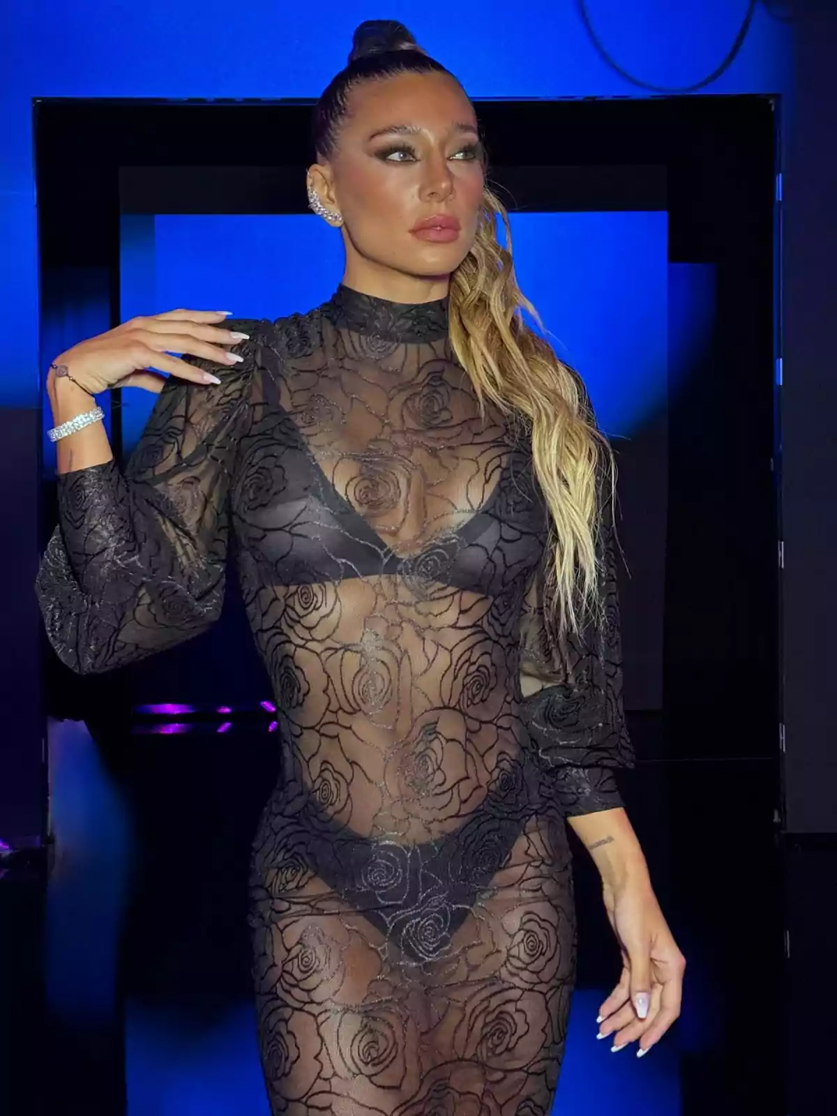Woman in a sheer black lace dress with a rose pattern posing in front of a blue background Woman in a sheer black lace dress with a rose pattern posing in front of a blue background