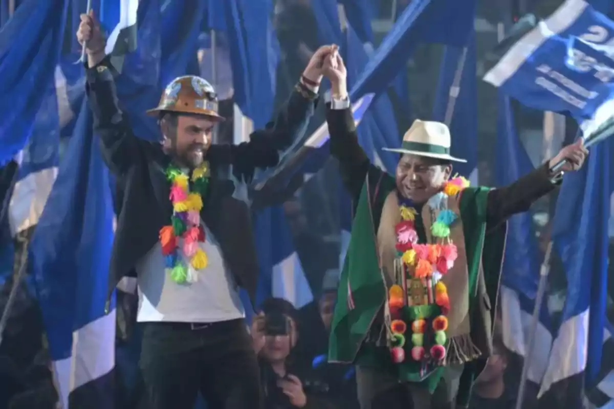 Dos hombres sonrientes con sombreros y guirnaldas de colores levantan las manos en señal de celebración mientras ondean banderas azules en un evento multitudinario
