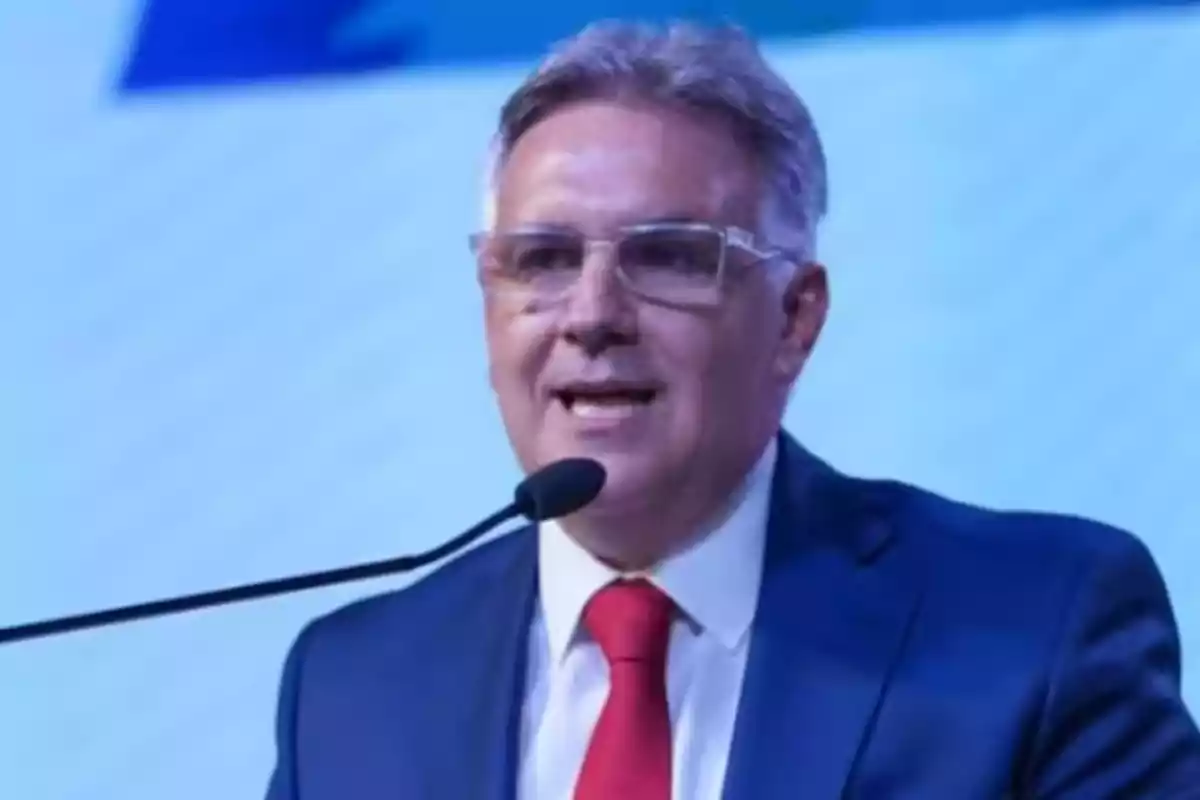 Hombre de cabello canoso y gafas hablando frente a un micrófono con fondo azul claro, viste traje azul y corbata roja