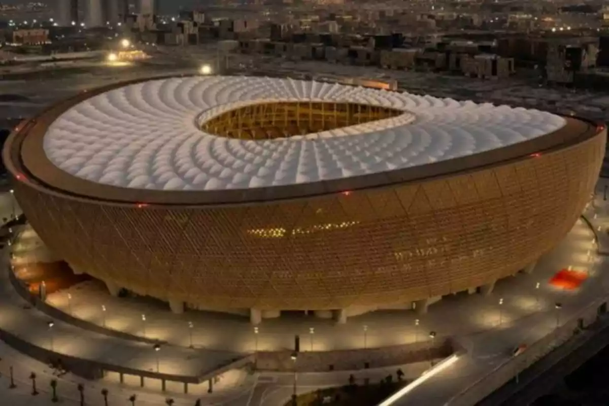 Vista nocturna de un estadio moderno con iluminación dorada y techo blanco en forma ovalada Vista nocturna de un estadio moderno con iluminación dorada y techo blanco en forma ovalada
