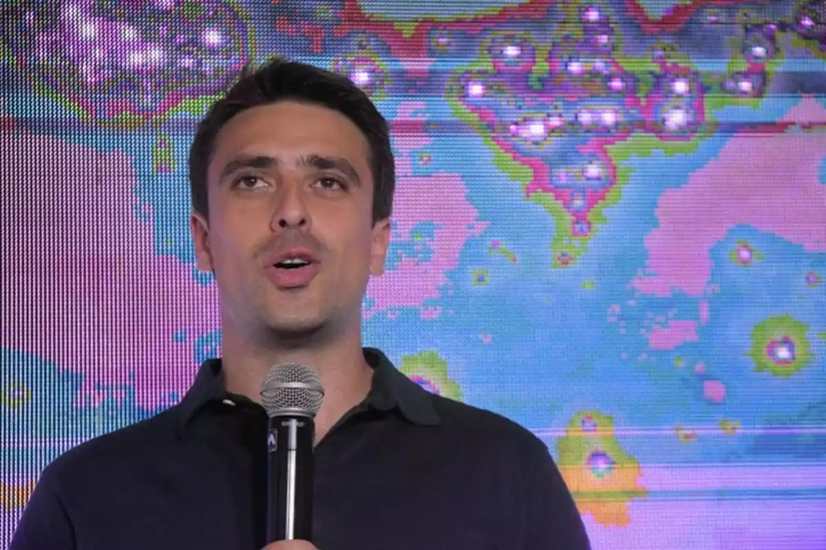 Hombre joven hablando frente a un micrófono con una pantalla colorida de fondo que muestra un mapa o gráfico abstracto Hombre joven hablando frente a un micrófono con una pantalla colorida de fondo que muestra un mapa o gráfico abstracto