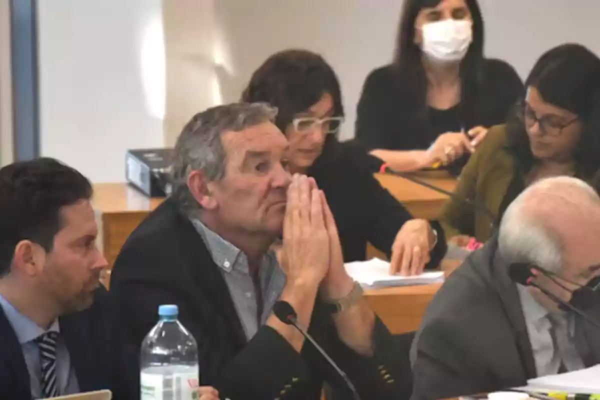 Older man with his hands clasped in a thoughtful manner, surrounded by other people in a meeting room Older man with his hands clasped in a thoughtful manner, surrounded by other people in a meeting room