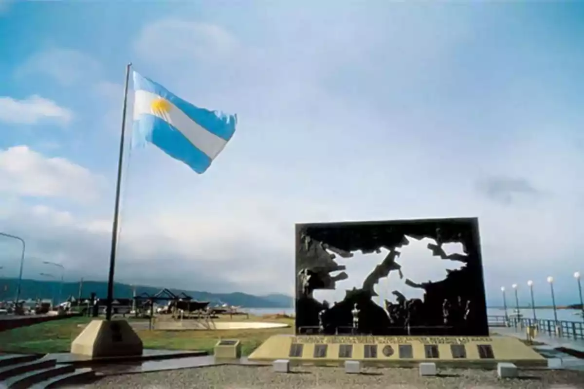 Bandera de Argentina ondeando junto a un monumento con la silueta de las Islas Malvinas en un espacio al aire libre