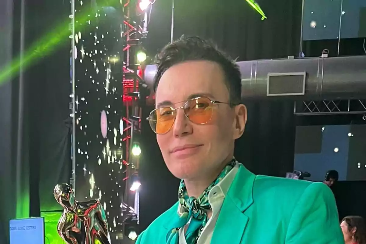 Person wearing orange sunglasses and a sea green jacket posing at an event with lights and a decorative star background