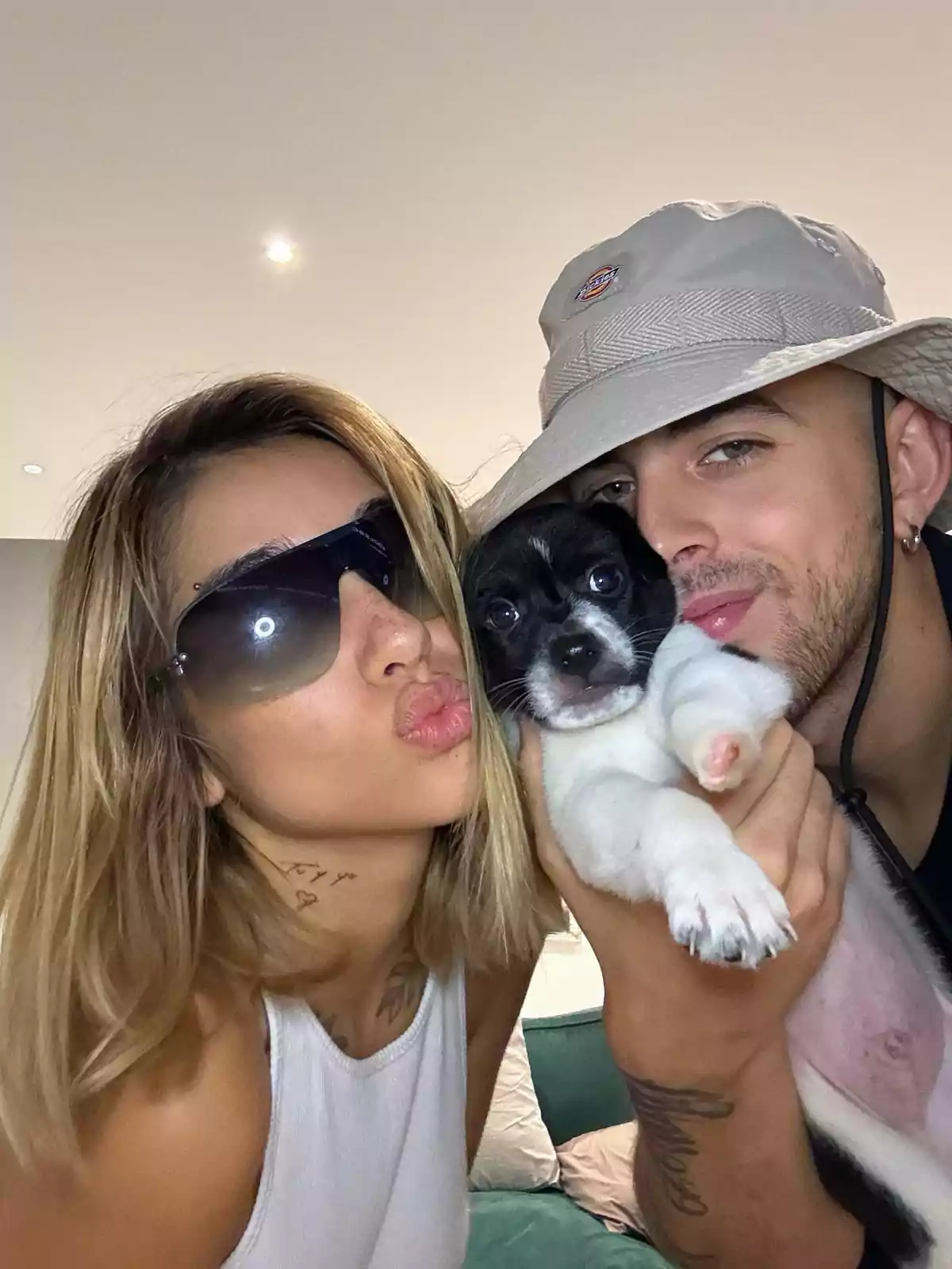 A couple smiles while holding a black and white puppy; the woman is wearing dark sunglasses and the man a light-colored hat. A couple smiles while holding a black and white puppy; the woman is wearing dark sunglasses and the man a light-colored hat.