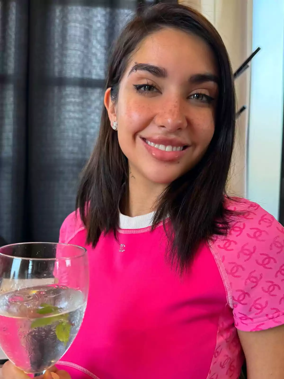 Young person with straight dark hair smiles while holding a glass with a drink and mint leaves, wearing a pink t-jersey with logo details and standing in front of a dark curtain. Young person with straight dark hair smiles while holding a glass with a drink and mint leaves, wearing a pink t-jersey with logo details and standing in front of a dark curtain.