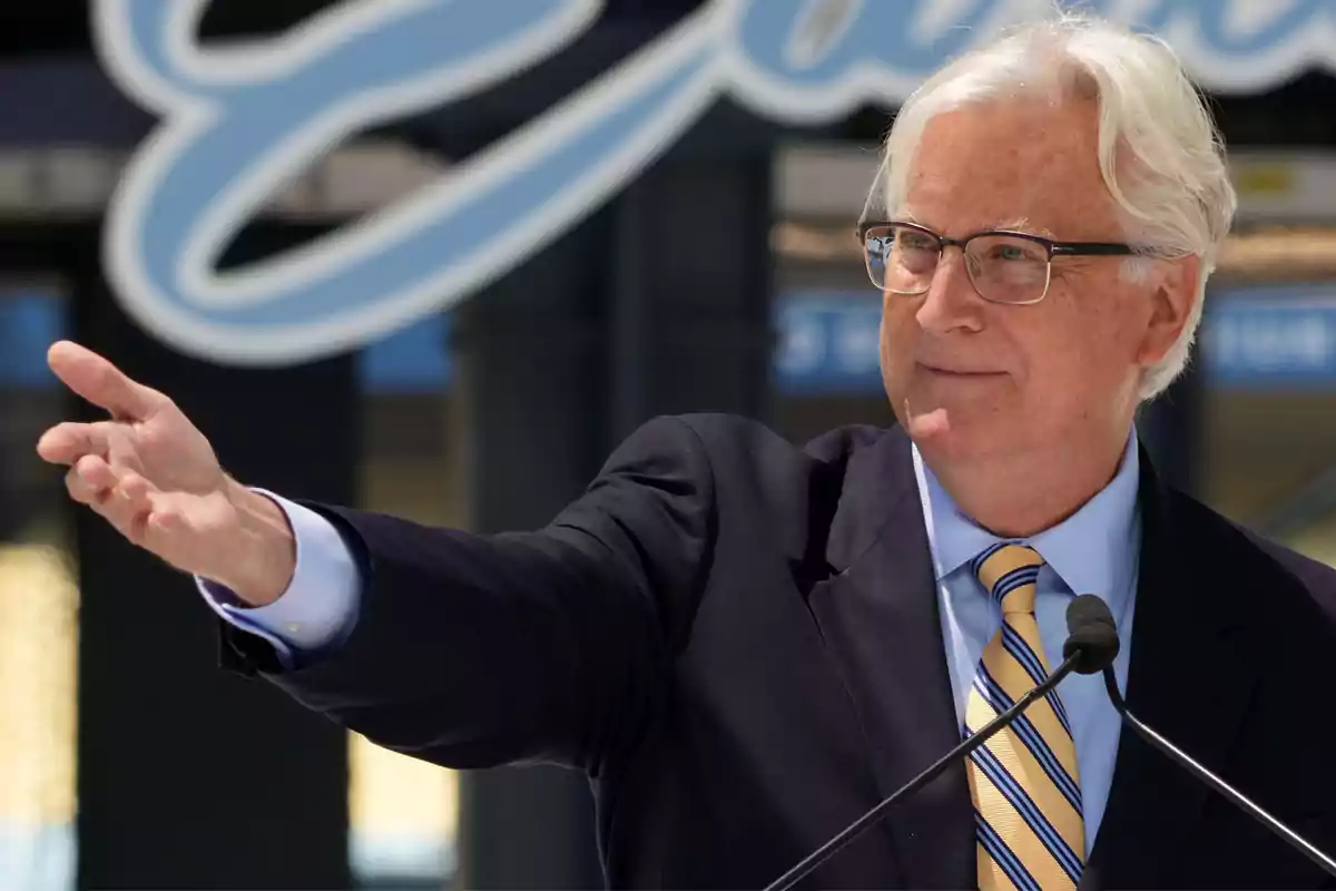 Gray-haired man with glasses wearing a dark suit and striped tie, speaking in front of a microphone and extending a hand Gray-haired man with glasses wearing a dark suit and striped tie, speaking in front of a microphone and extending a hand
