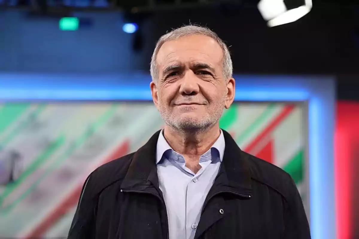 Gray-haired man with a short beard wearing a light blue jersey and dark jacket posing in front of a colorful background in a television studio Gray-haired man with a short beard wearing a light blue jersey and dark jacket posing in front of a colorful background in a television studio