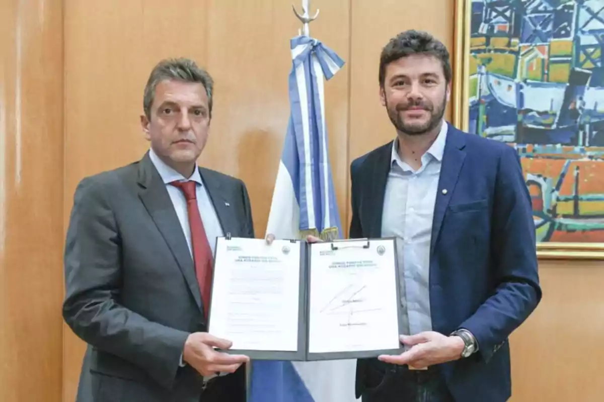 Dos hombres de traje posan juntos en una oficina sosteniendo un documento abierto, detrás de ellos hay una bandera y un cuadro colorido en la pared.