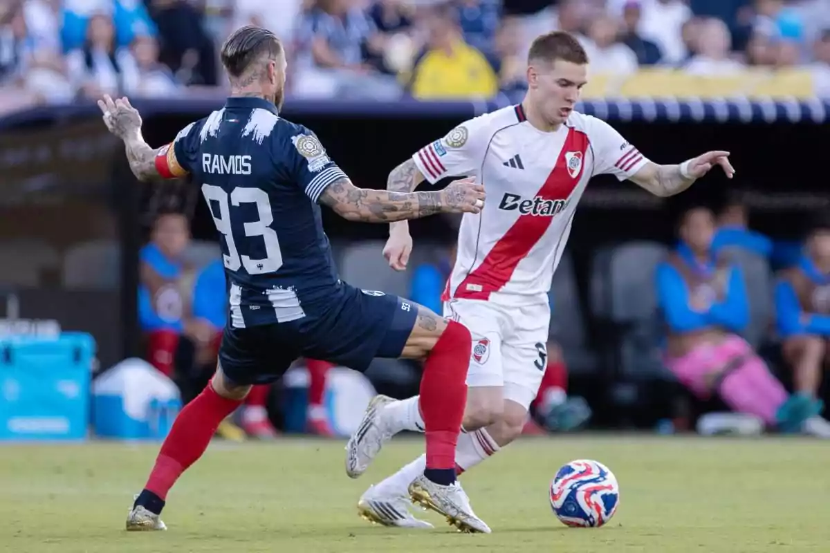 Dos futbolistas disputan el balón durante un partido mientras el público observa desde las gradas Dos futbolistas disputan el balón durante un partido mientras el público observa desde las gradas