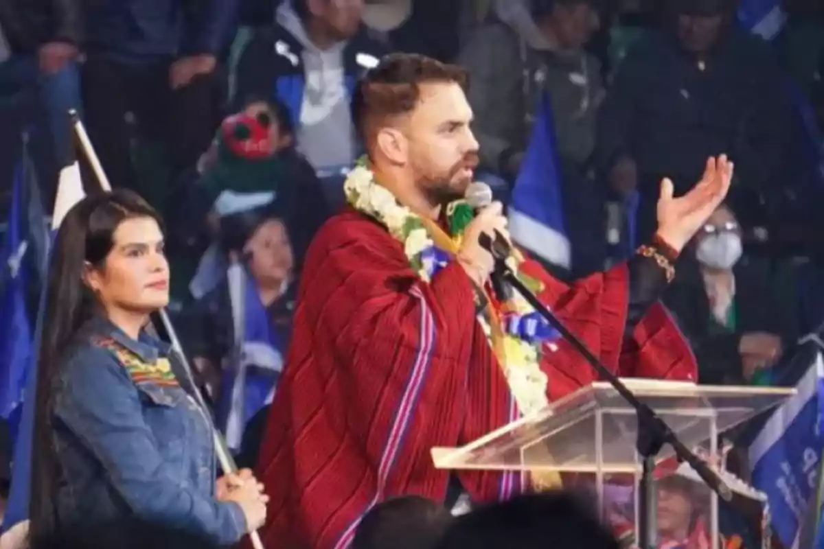 Un hombre con poncho rojo y guirnalda de flores habla en un micrófono desde un podio mientras una mujer de cabello largo y chaqueta de mezclilla lo acompaña, al fondo se observa un público con banderas.