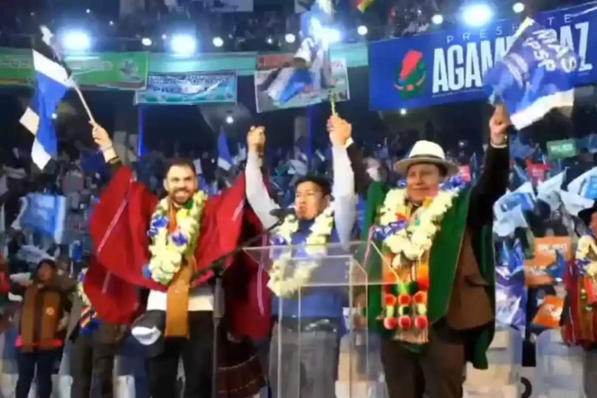 Tres personas en un escenario levantan los brazos y ondean banderas azules mientras llevan guirnaldas de flores y ponchos típicos, rodeados de una multitud en un evento político.