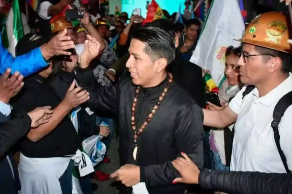 Un hombre con collar de cuentas camina entre una multitud de personas que lo saludan y lo rodean, algunas llevan cascos y banderas Un hombre con collar de cuentas camina entre una multitud de personas que lo saludan y lo rodean, algunas llevan cascos y banderas