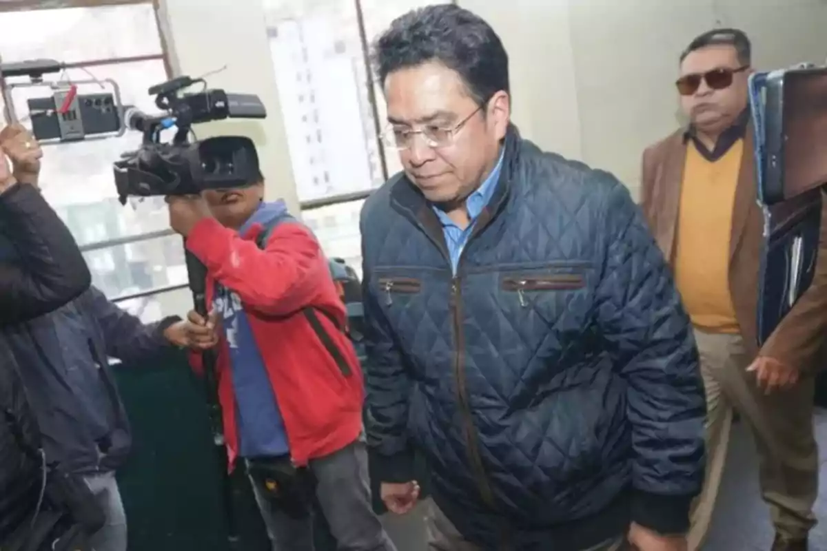 A man in a blue jacket walks inside a building while being filmed by a cameraman and accompanied by other people.