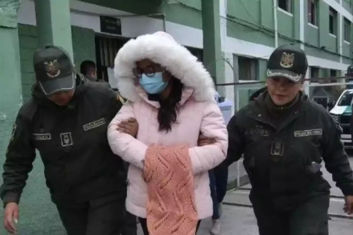 Woman in a pink coat and face mask is escorted by two police officers in dark uniforms in front of a green building. Woman in a pink coat and face mask is escorted by two police officers in dark uniforms in front of a green building.