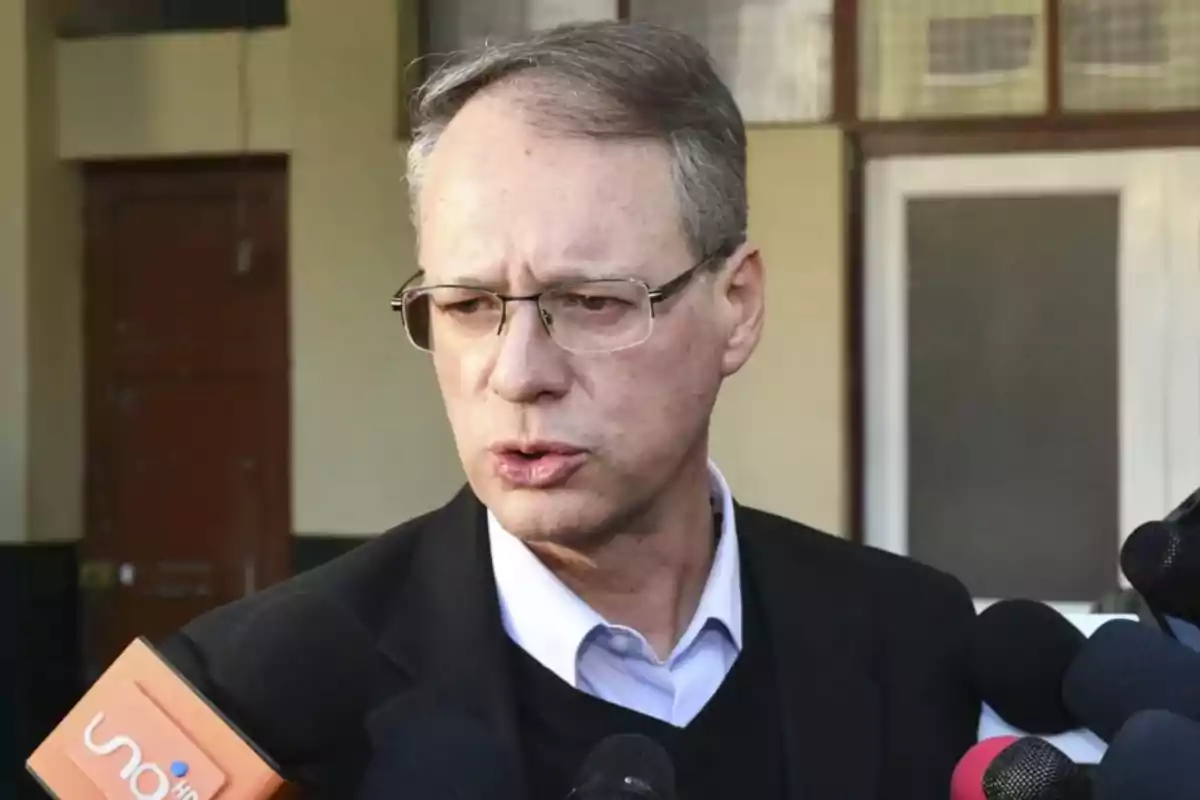 Gray-haired man with glasses speaking in front of several microphones during an outdoor interview