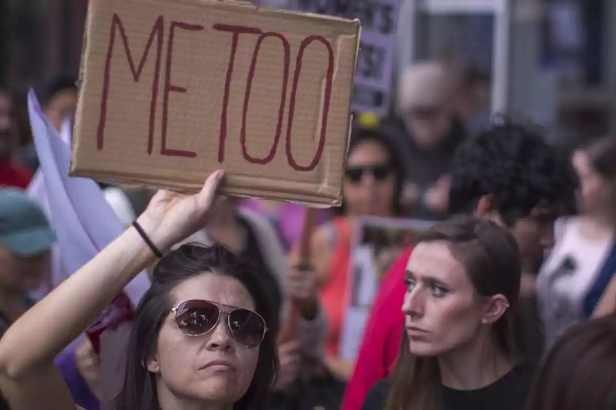 Mujer con gafas de sol sostiene un cartel de cartón con la palabra METOO durante una manifestación rodeada de otras personas Mujer con gafas de sol sostiene un cartel de cartón con la palabra METOO durante una manifestación rodeada de otras personas