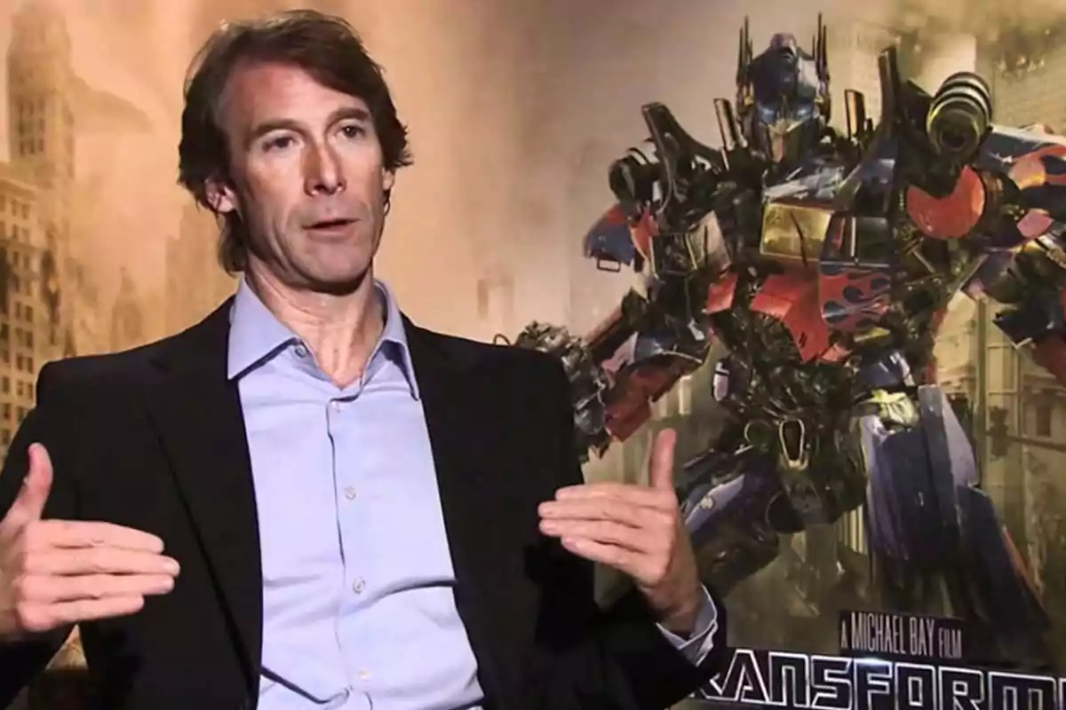 Hombre de cabello castaño con saco negro y camisa azul claro gesticulando con las manos mientras está sentado frente a un póster de la película Transformers donde aparece un robot grande Hombre de cabello castaño con saco negro y camisa azul claro gesticulando con las manos mientras está sentado frente a un póster de la película Transformers donde aparece un robot grande
