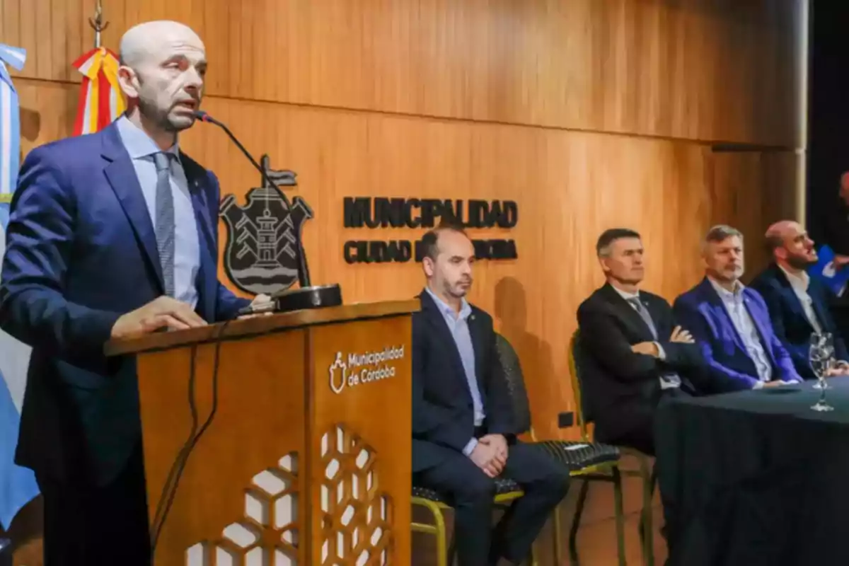 A man in a suit speaks at a podium during an official event at the Municipality of Córdoba while four other people listen to him seated beside him. A man in a suit speaks at a podium during an official event at the Municipality of Córdoba while four other people listen to him seated beside him.