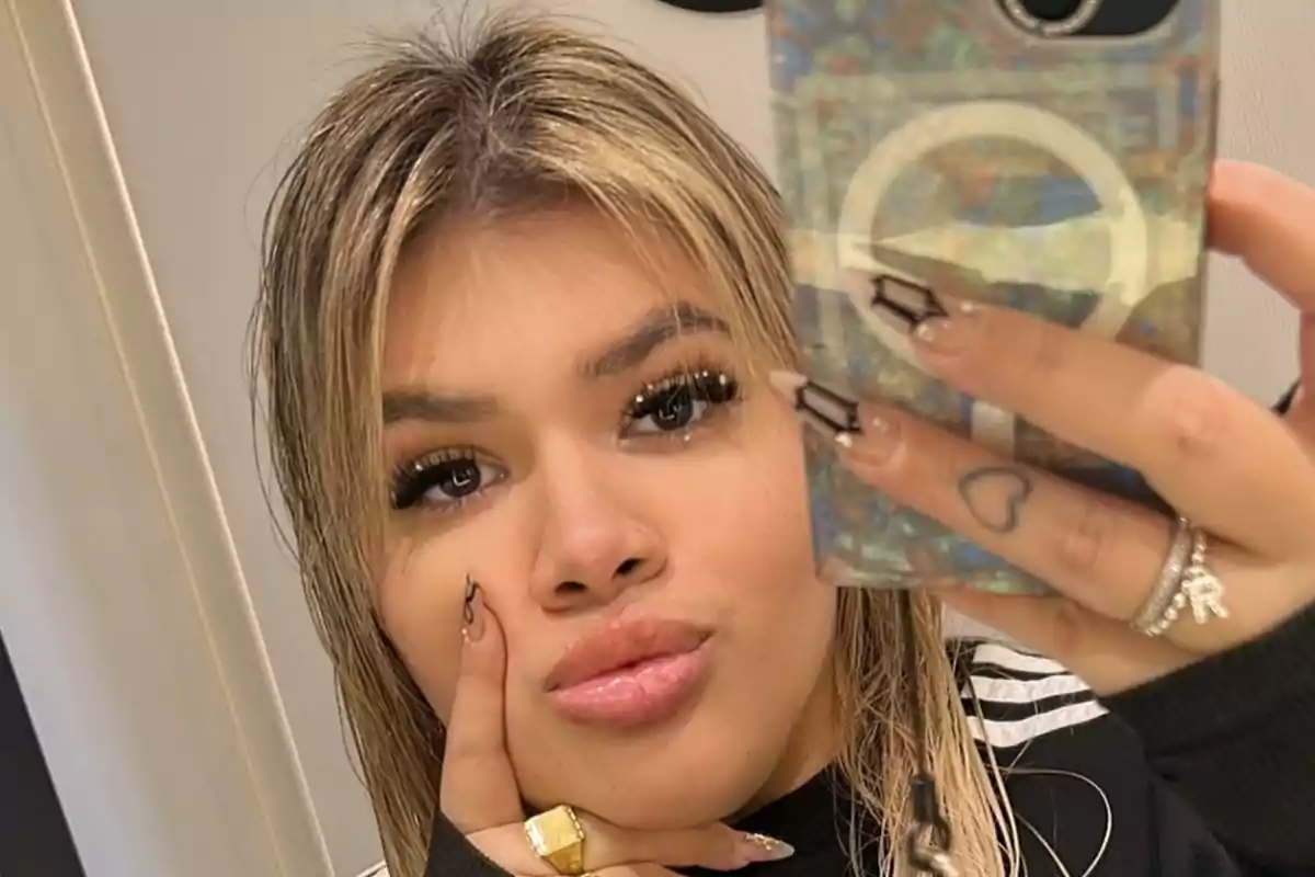 Young person taking a selfie in front of the mirror with blonde hair and decorated nails, wearing a gold ring and a black sweater with white stripes