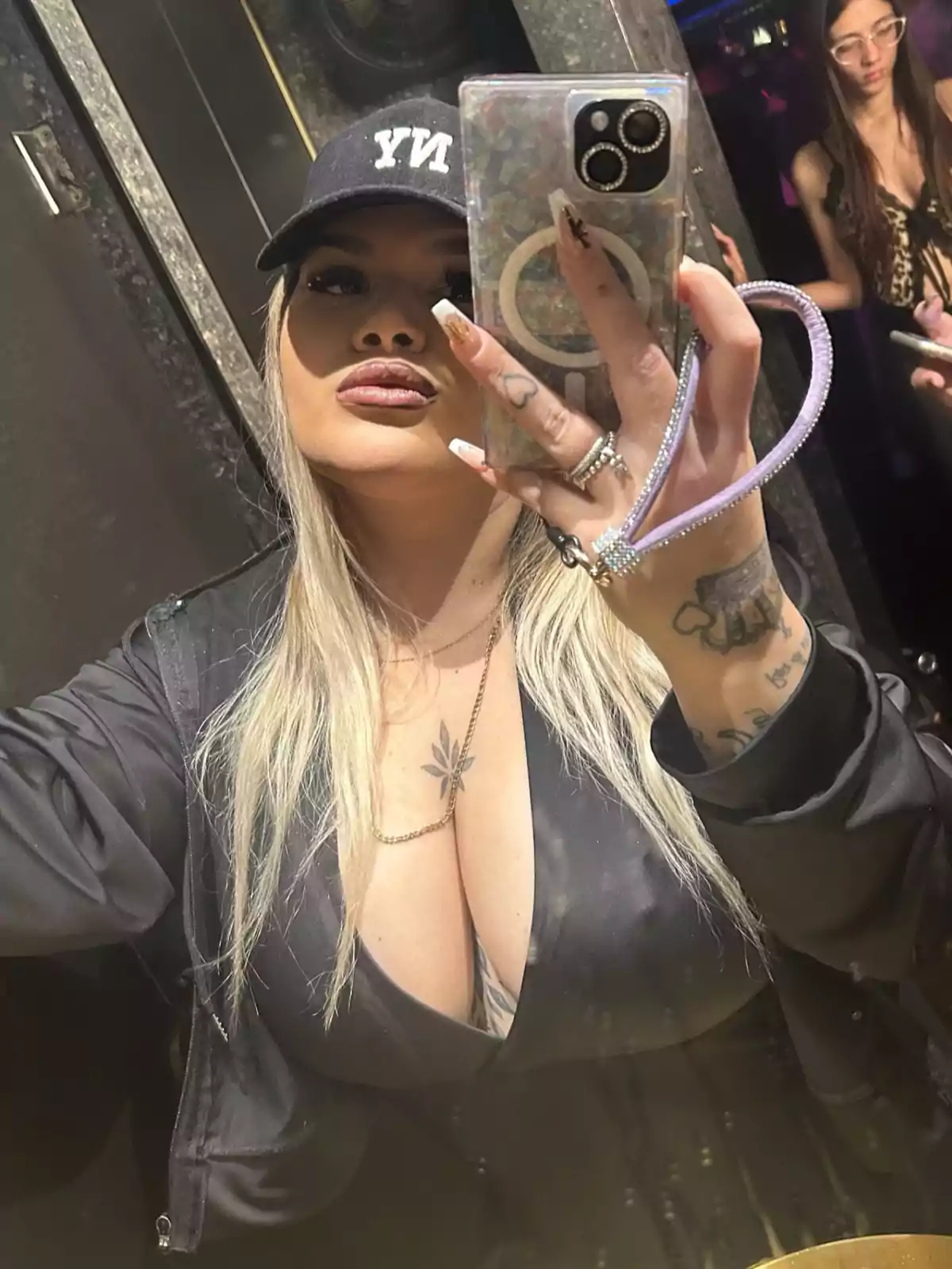 Mujer rubia con gorra negra que dice NY tomándose una selfie frente a un espejo en un lugar cerrado, lleva uñas largas decoradas y un tatuaje visible en el pecho, al fondo se ve otra persona Mujer rubia con gorra negra que dice NY tomándose una selfie frente a un espejo en un lugar cerrado, lleva uñas largas decoradas y un tatuaje visible en el pecho, al fondo se ve otra persona