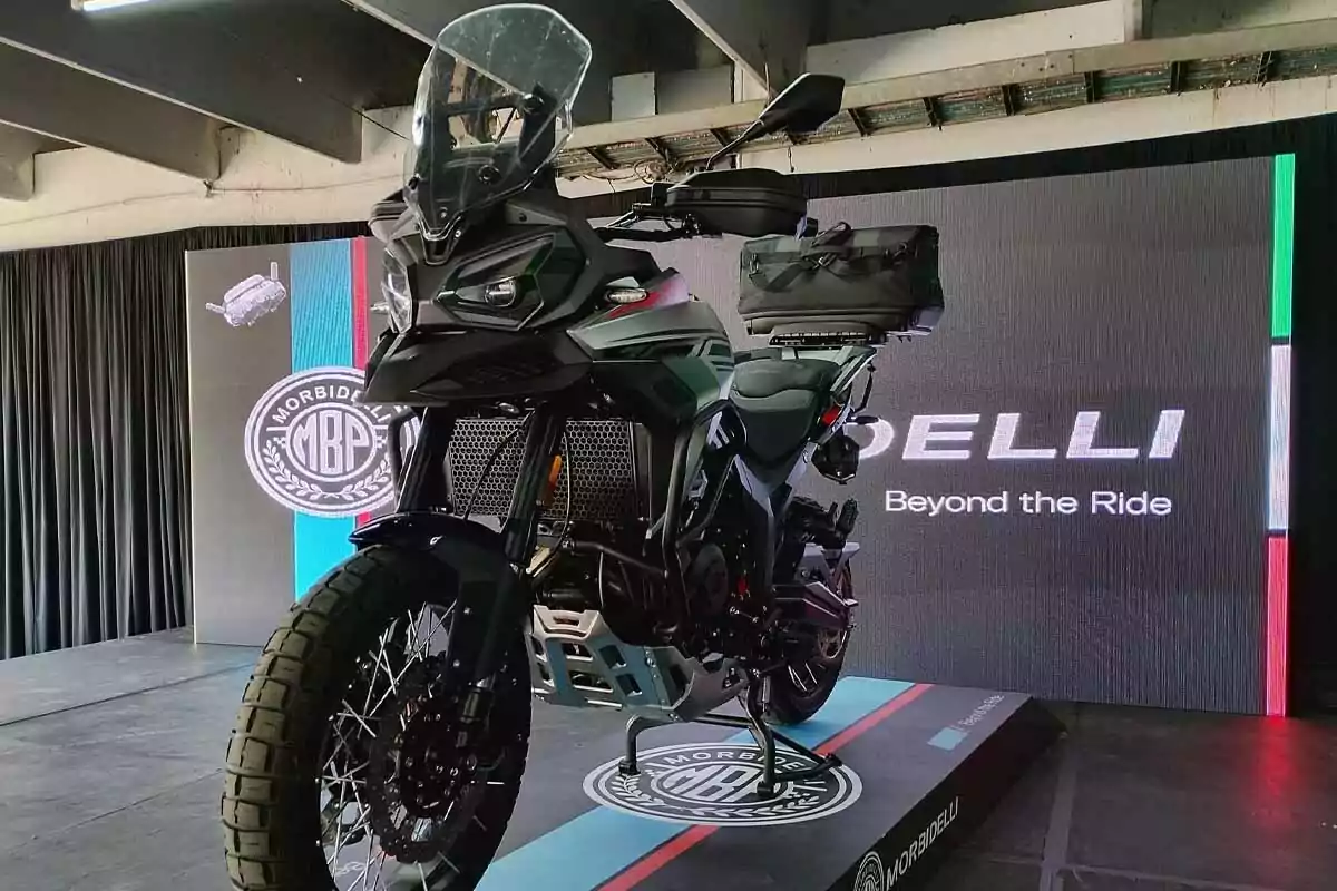 Motocicleta de aventura exhibida en una plataforma durante un evento de presentación con un fondo de pantalla que muestra el logo de Morbidelli y la frase Beyond the Ride Motocicleta de aventura exhibida en una plataforma durante un evento de presentación con un fondo de pantalla que muestra el logo de Morbidelli y la frase Beyond the Ride