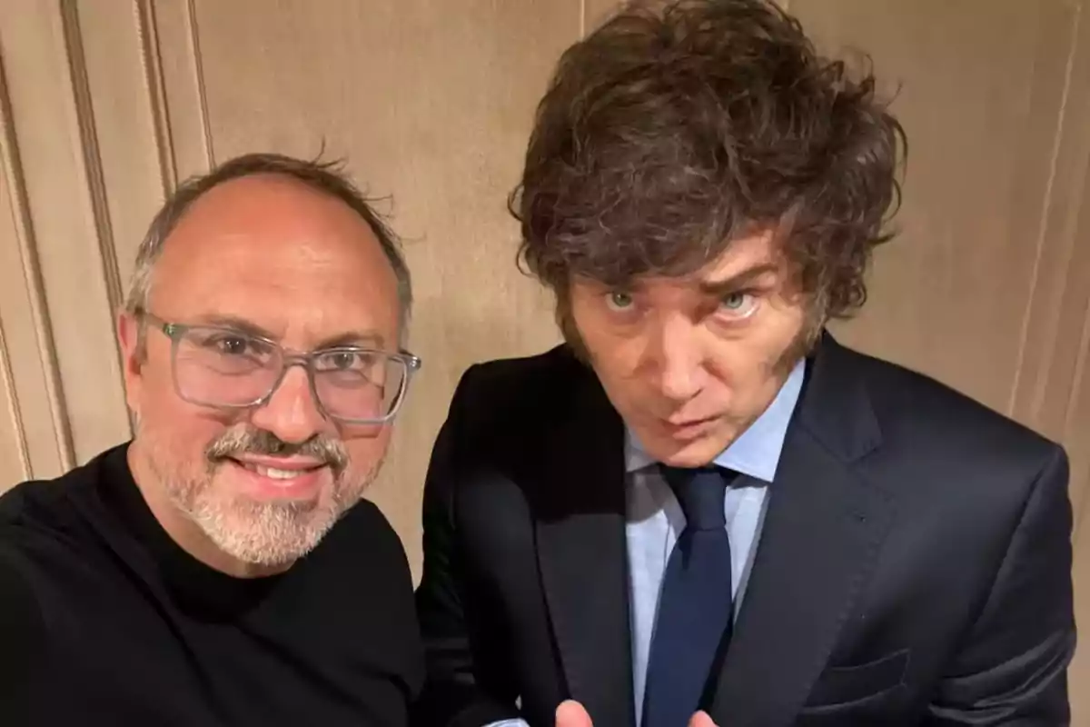 Dos hombres posan juntos para una selfie, uno lleva gafas y camiseta negra, el otro viste traje oscuro y corbata azul Dos hombres posan juntos para una selfie, uno lleva gafas y camiseta negra, el otro viste traje oscuro y corbata azul