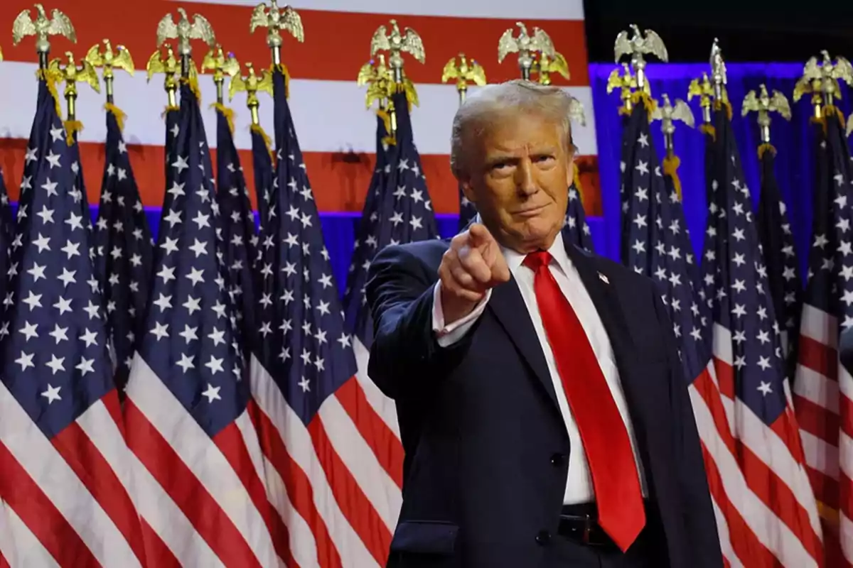 Hombre de traje señalando al frente con varias banderas de Estados Unidos de fondo