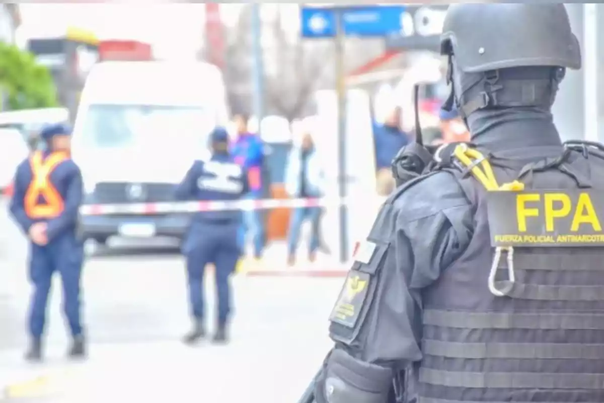 Agente de la Fuerza Policial Antinarcotráfico con chaleco y casco observando un operativo en la vía pública donde hay otros policías y una cinta de seguridad Agente de la Fuerza Policial Antinarcotráfico con chaleco y casco observando un operativo en la vía pública donde hay otros policías y una cinta de seguridad
