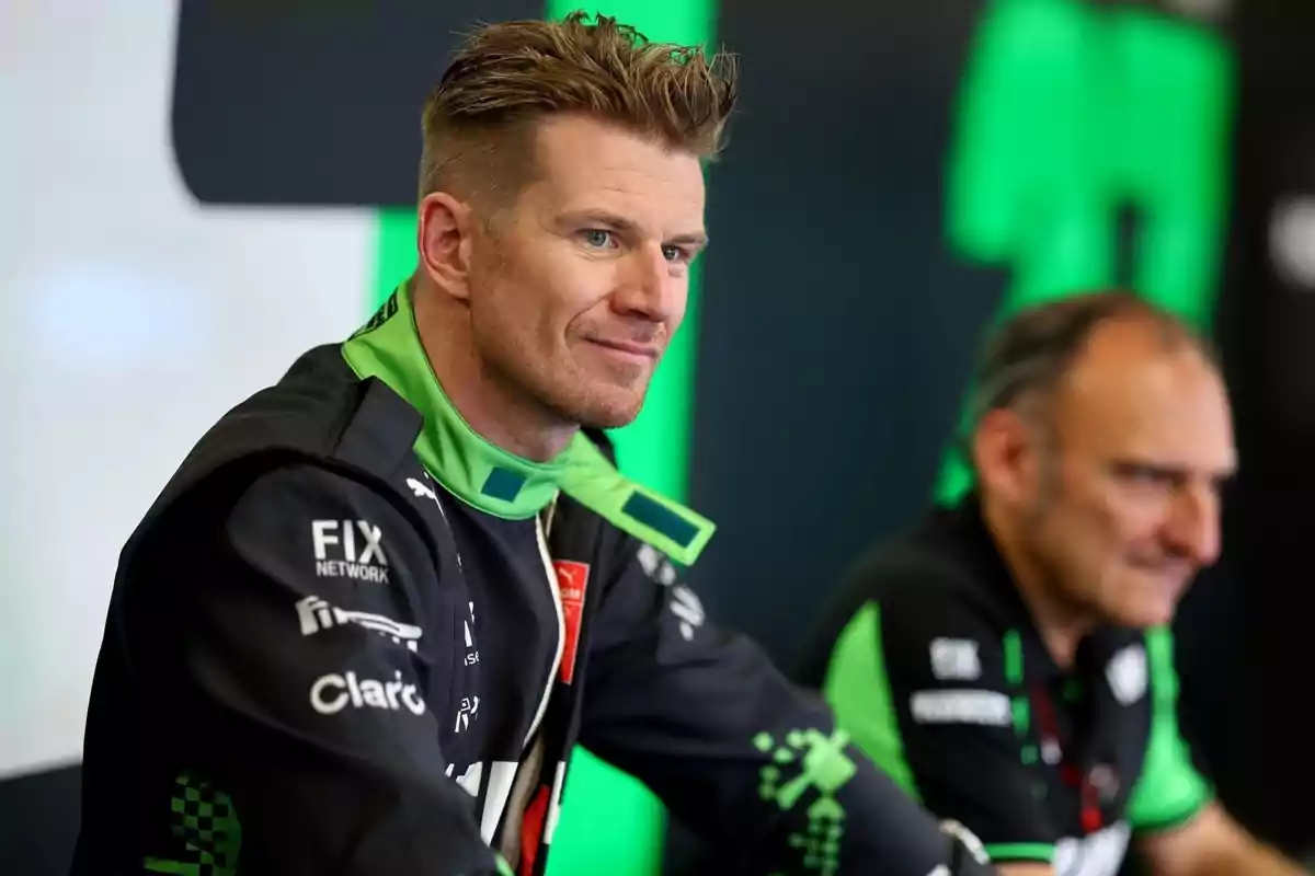 Piloto de automovilismo con uniforme negro y verde sentado junto a otra persona en un entorno de equipo de carreras Piloto de automovilismo con uniforme negro y verde sentado junto a otra persona en un entorno de equipo de carreras