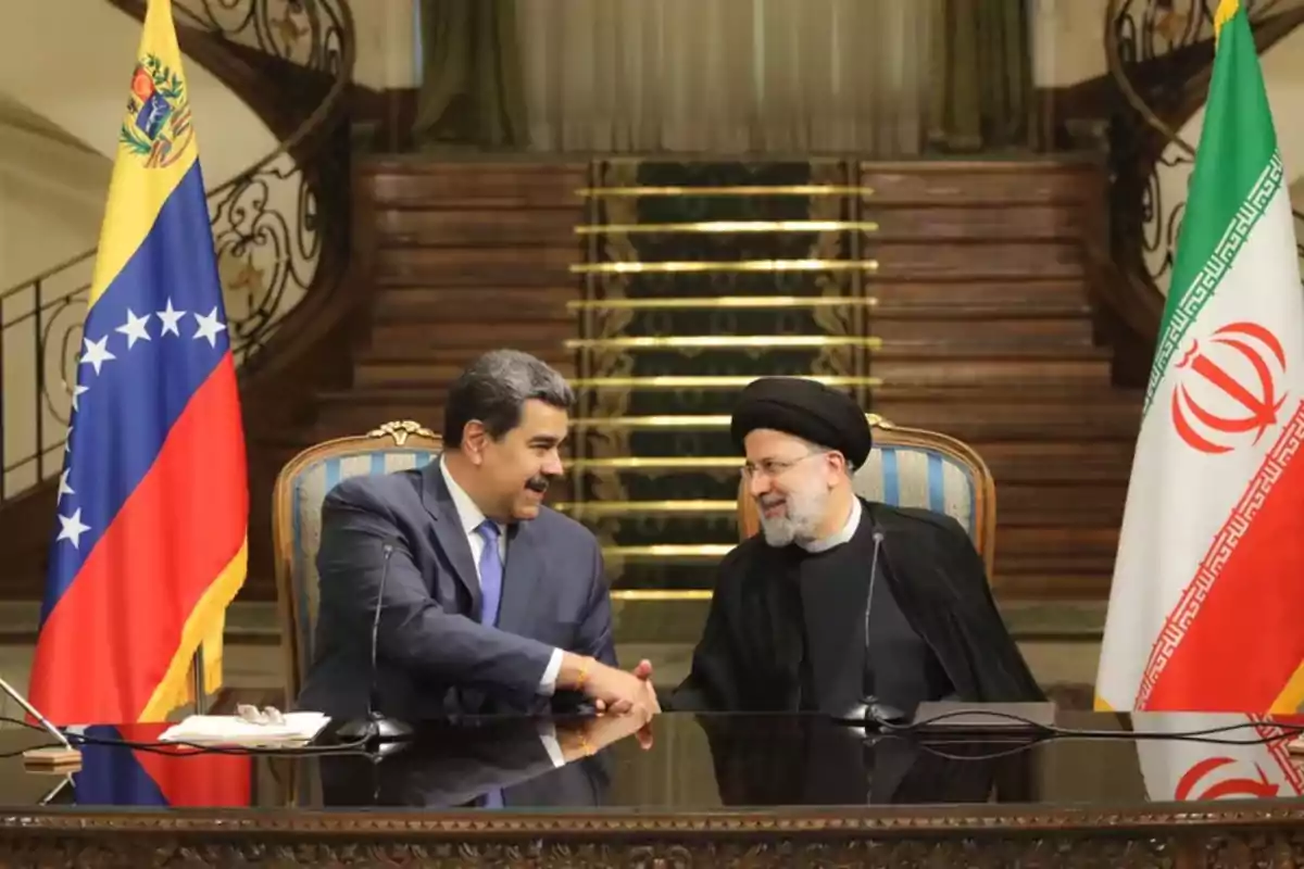Two men sitting at a table shake hands and smile, with the flags of Venezuela and Iran on either side and a staircase in the background. Two men sitting at a table shake hands and smile, with the flags of Venezuela and Iran on either side and a staircase in the background.