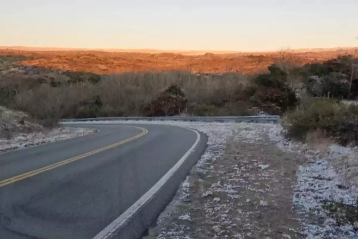Curvy road surrounded by vegetation with frost or light snow on the ground and sunlit hills in the background