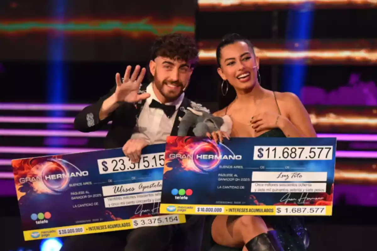 Dos personas sonrientes sentadas en un escenario sostienen cheques gigantes de premio del programa Gran Hermano, una de ellas saluda con la mano y la otra sostiene un peluche. Dos personas sonrientes sentadas en un escenario sostienen cheques gigantes de premio del programa Gran Hermano, una de ellas saluda con la mano y la otra sostiene un peluche.