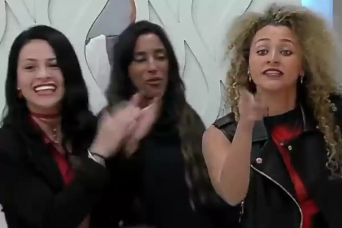 Three women are indoors; one of them is smiling and clapping, another is in the center with a neutral expression, and the third is pointing forward with an emphatic gesture. Three women are indoors; one of them is smiling and clapping, another is in the center with a neutral expression, and the third is pointing forward with an emphatic gesture.
